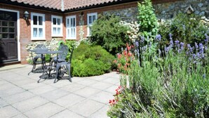 A brick house with a tiled roof, a patio with a table and chairs, and a garden with lavender and red flowers. - 2 Bed in Weasenham (oc-kt103) (Weasenham)