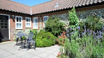 A brick house with a tiled roof, a patio with a table and chairs, and a garden with lavender and red flowers.
