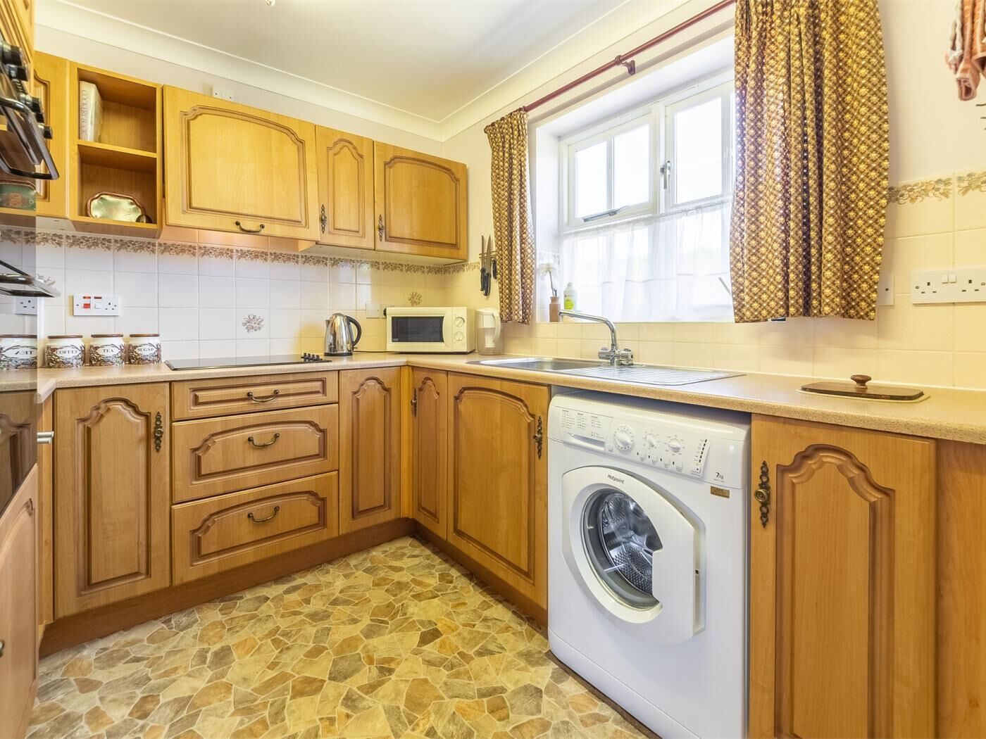 A compact kitchen with wooden cabinets, a washing machine, a microwave, and a window with curtains.