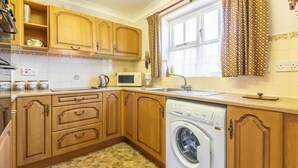 A compact kitchen with wooden cabinets, a washing machine, a microwave, and a window with curtains. - 2 Bed in Weasenham (oc-kt103) (Weasenham)