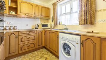 A compact kitchen with wooden cabinets, a washing machine, a microwave, and a window with curtains.