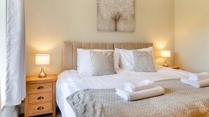 Een net opgemaakt bed met witte lakens en een beige deken, twee nachtkastjes met lampen, een houten nachtkastje met lades, en een schilderij van een boom aan de muur.