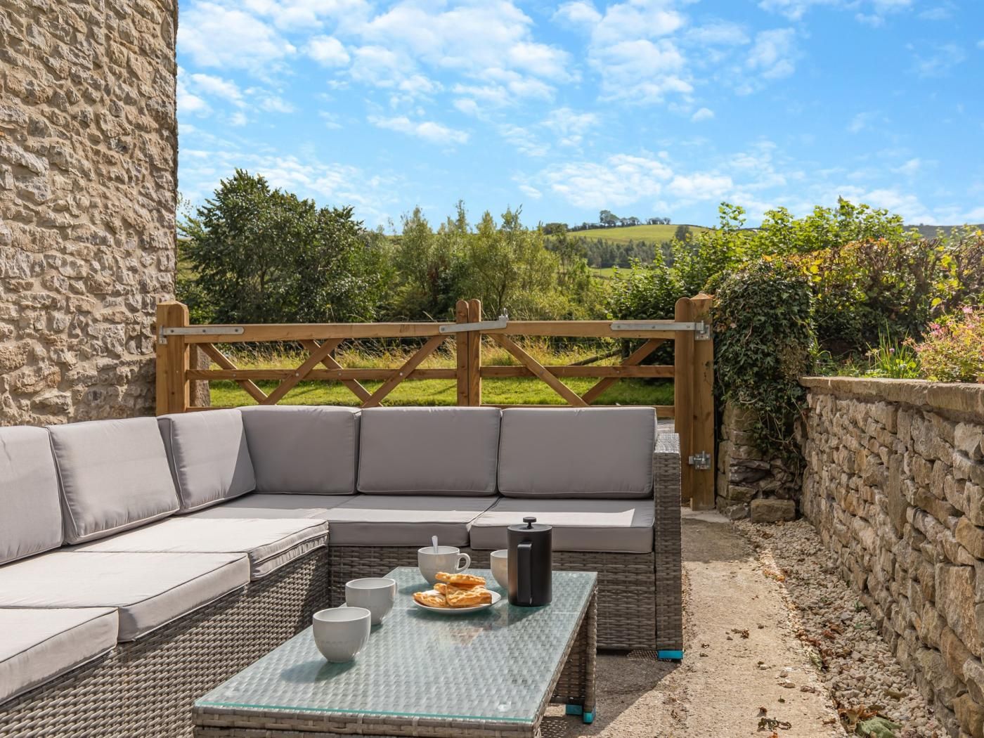 An outdoor seating area with a stone wall, a wooden gate, and a view of a green landscape.