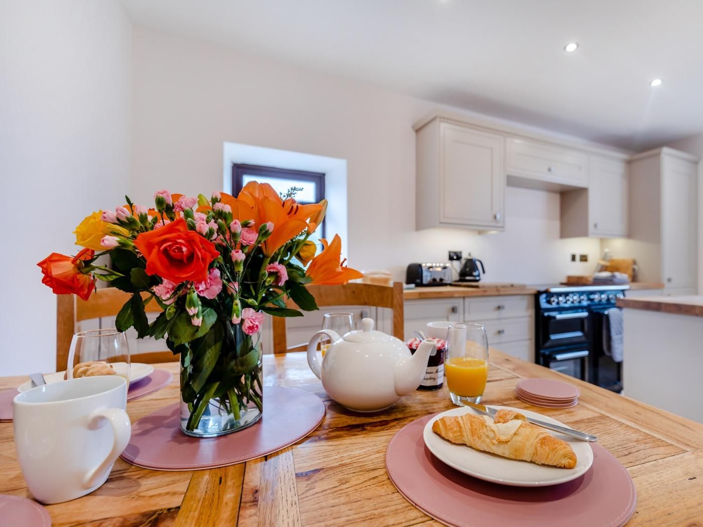 Une table de petit-déjeuner avec un vase de fleurs, une théière, une tasse, une assiette avec un croissant et un verre de jus d’orange.