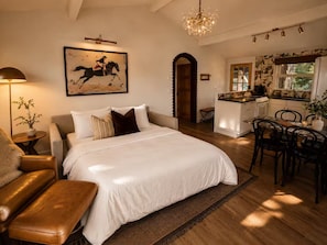 A bedroom with a large bed, a sitting area with a bench, a dining area with chairs, a kitchen with a stove, and a chandelier. - Magical Retreat Over Morse Creek with Hot Tub (Port Angeles)
