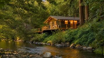 A cabin with a deck overlooking a river, surrounded by dense forest.