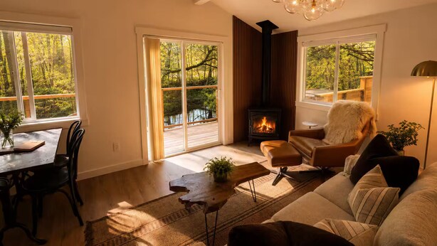 Ein gemütliches Wohnzimmer mit Kamin, einer Couch, einem Esstisch und großen Fenstern mit Blick auf Bäume.