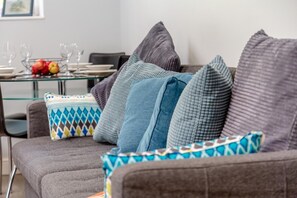Ein modernes Wohnzimmer mit einem grauen Sofa, vielen Kissen, einem Glastisch mit Früchten und einem Essbereich im Hintergrund.