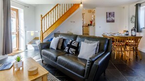 A modern living room with a black leather sofa, wooden staircase, and a dining area with wooden chairs. - 1 Bed in Lydney (oc-43724) (Lydney)