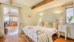 Een slaapkamer met een bed, nachtkastjes, een raam met bloemen en uitzicht op een keuken en eetgedeelte.