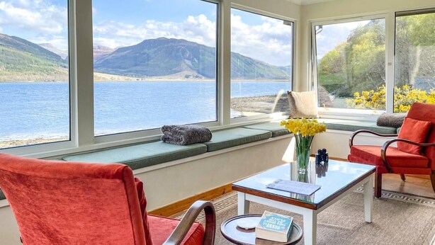 Ein Zimmer mit Blick auf einen See und Berge, ein roter Sessel, ein Tisch mit einem Buch und eine Vase mit gelben Blumen.