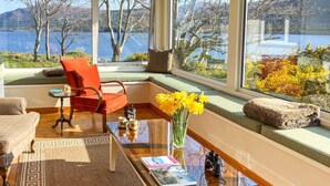 Ein gemütliches Zimmer mit Seeblick, ein roter Sessel, ein Glastisch und eine Vase mit gelben Blumen.