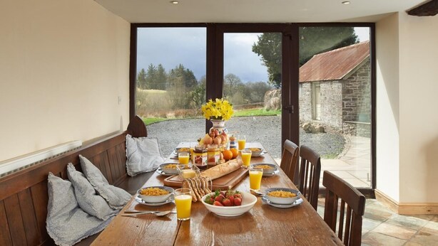Mesa posta para o pequeno-almoço, com almofadas num banco, e vista para um edifício de pedra e vegetação.