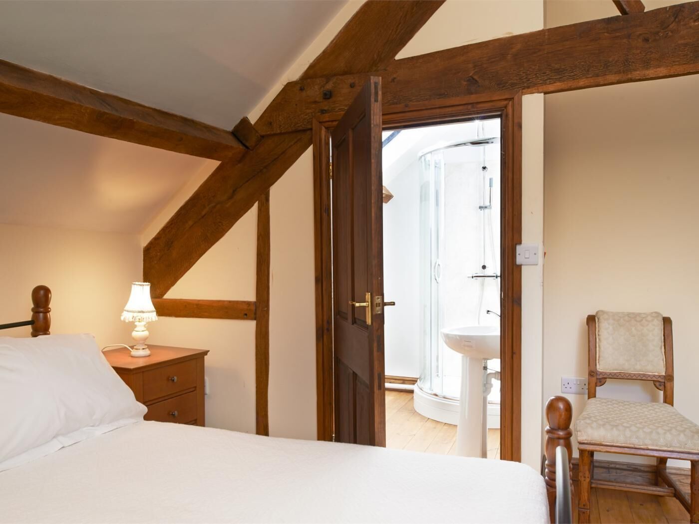 A bedroom with a bed, bedside table, lamp, wooden beams, a door leading to a bathroom, and a chair.