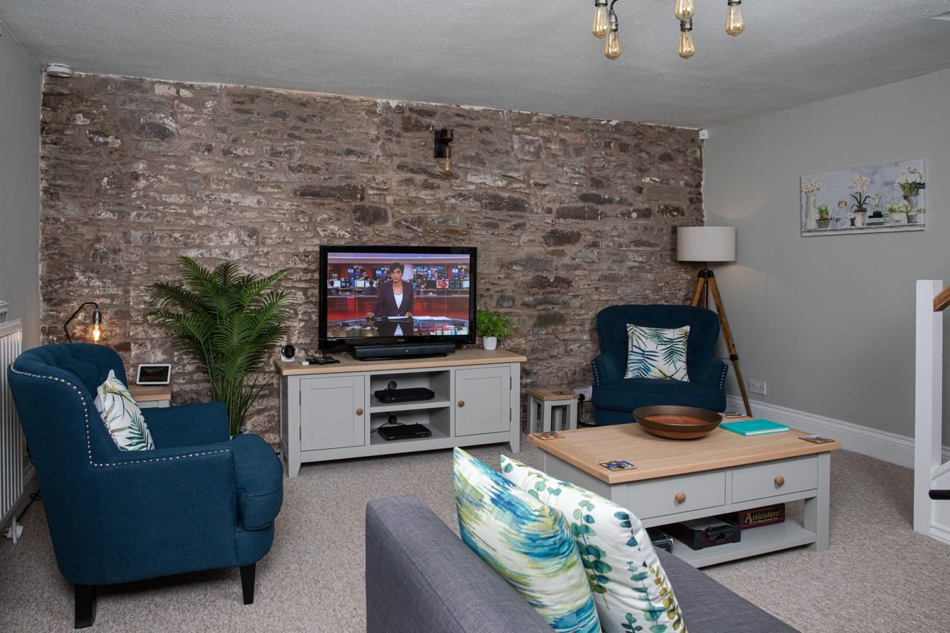 A living room with a stone wall, a television, a blue armchair, a grey sofa, a coffee table, and a lamp.