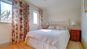 Ein Schlafzimmer mit Holzboden, einem weißen Bett, blumigen Vorhängen, einem Nachttisch mit Lampe und einem Fenster.