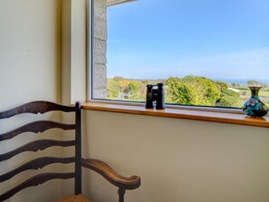 Ein Fenster mit Blick auf Grün und ein Gewässer, ein Holzstuhl und eine Vase auf dem Fensterbrett.