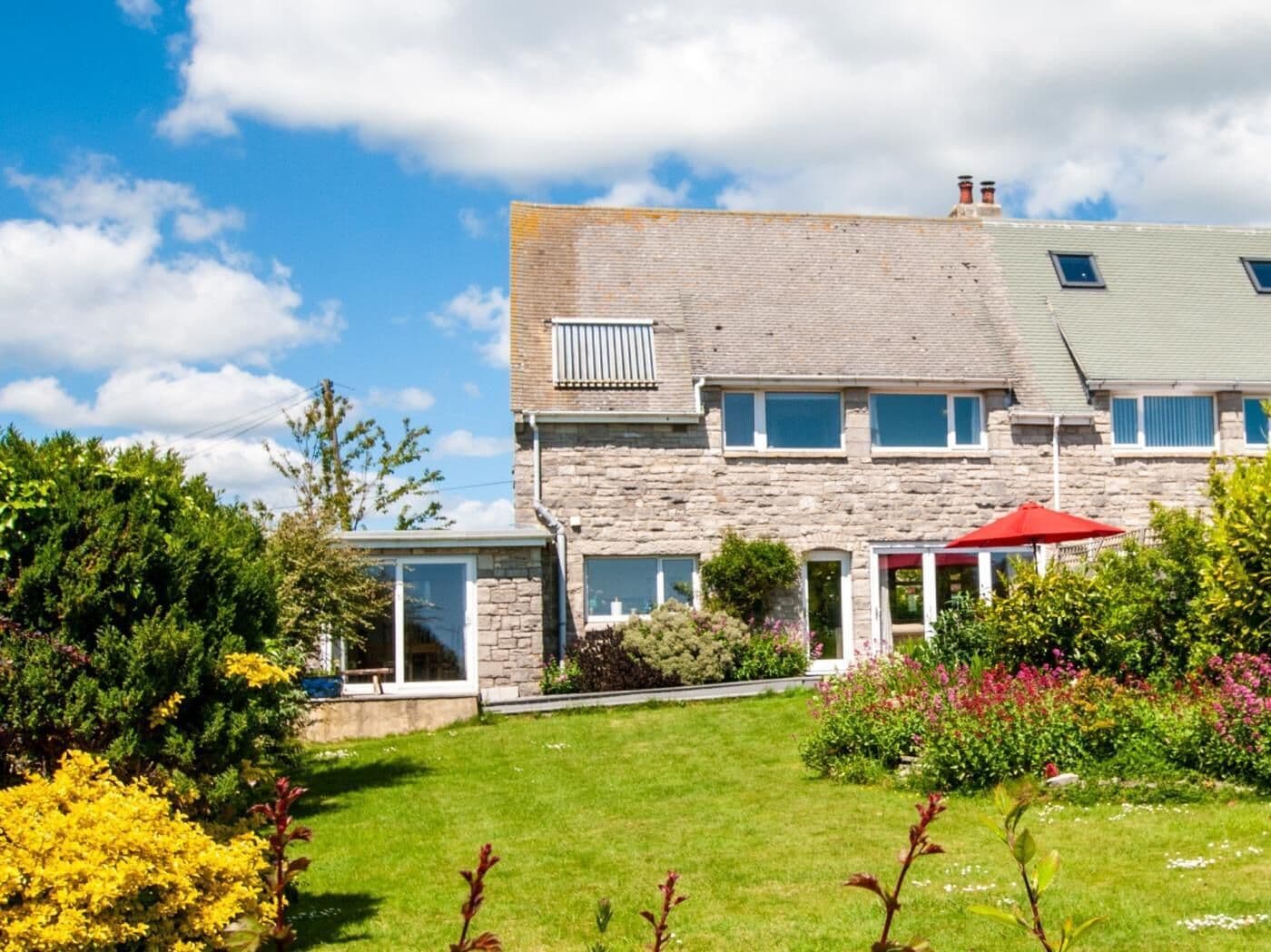 Ein zweistöckiges Steinhaus mit einem grünen Dach, einer überdachten Terrasse und einem Garten mit verschiedenen Pflanzen und Blumen.