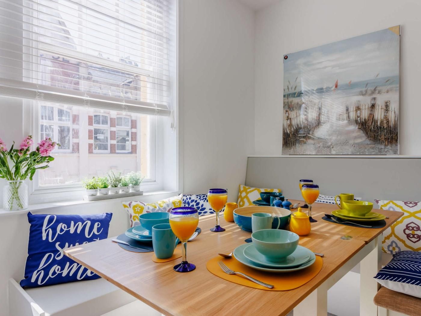 En matplats med ett träbord dukat för frukost, med färgglada tallrikar och glas med apelsinjuice.