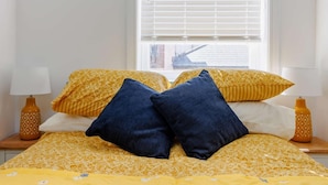 Ein Bett mit gelbem, gemusterten Bettzeug, zwei dunkelblaue Kissen und ein Fenster mit Jalousien.