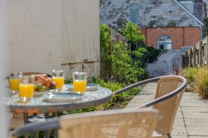 An outdoor dining area with a table set for two, glasses of orange juice, and a view of a brick building in the background. - 2 Bed in Padstow (oc-88178) (Padstow)