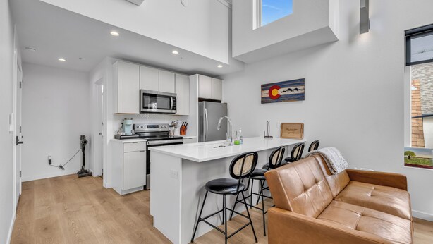 A modern kitchen with white cabinetry, a central island with bar stools, and a brown leather sofa in a bright, open space. - Rino Private Rooftop  - Art District Location. Walk To Everything! (Denver)