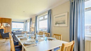 A dining area with a white table set for four, wooden chairs, and a view of the sea through large windows. - 4 Bed in Scourie (oc-ca399) (Scourie More)