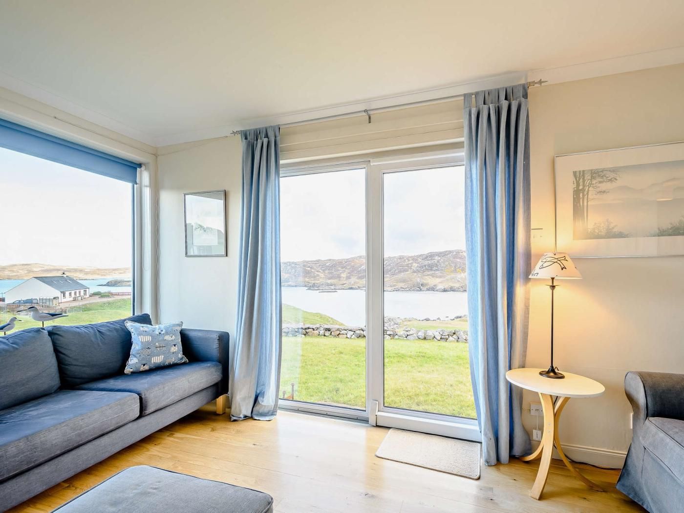 Een moderne woonkamer met een grijze bank, een klein rond tafeltje en uitzicht op een meer en heuvels.