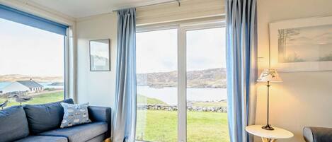 Een moderne woonkamer met een grijze bank, een klein rond tafeltje en uitzicht op een meer en heuvels.