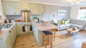 A modern kitchen with light green cabinets, a stainless steel range hood, and a built-in oven. There's a wooden bar stool and a small island with a basket. A gray sofa and a wooden coffee table are in the living area, with a window and a plant in the background. - 2 Bed in York (oc-81052) (Helmsley)