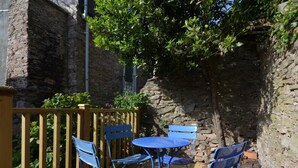 A stone building with a wooden deck, blue chairs, and a small round table. - 2 Bed in Brixham (oc-bx087) (Brixham)