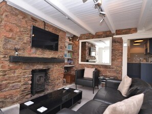 A living room with a stone fireplace, a black leather sofa, a flat-screen TV mounted on the wall, and a kitchen visible through an open doorway. - 2 Bed in Brixham (oc-bx087) (Brixham)