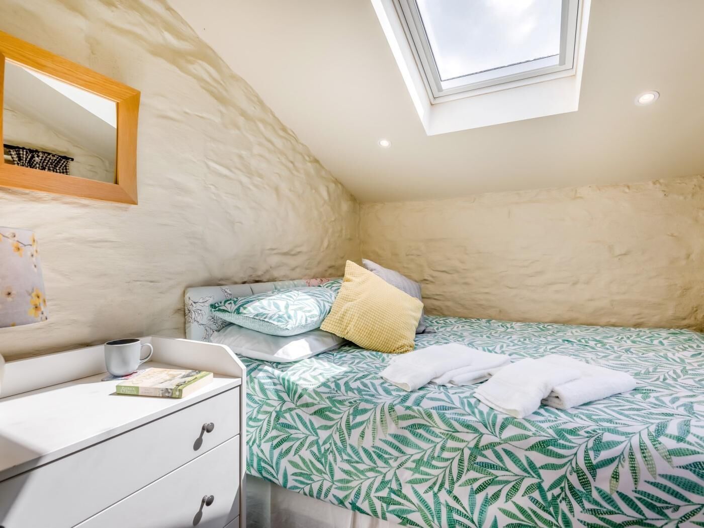Een gezellige slaapkamer met een schuine kap, een dakraam, een bed met een bedsprei met een bladmotief, een nachtkastje met een kopje en boek, en een zachte, geruite wand.