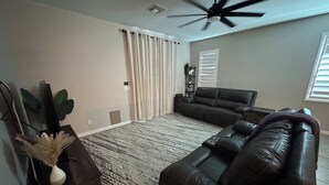 Un salon comprenant un canapé d’angle, un ventilateur de plafond, un tapis à motifs et une plante.