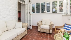 Une pièce avec un mur blanc à texture, un canapé blanc, un fauteuil en osier, une chaise en bois et une fenêtre donnant sur un paysage verdoyant.