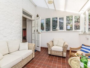 Ein Zimmer mit einer weißen Wand mit Struktur, einem weißen Sofa, einem Korbstuhl, einem Holzstuhl und einem Fenster mit Blick auf Grünflächen.