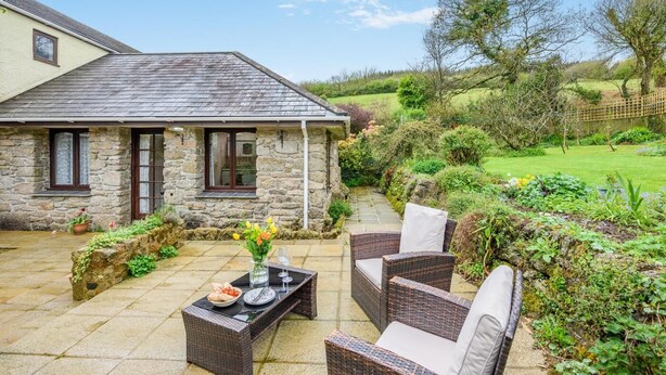 Une maison en pierre avec un patio carrelé, des meubles en osier et un jardin.