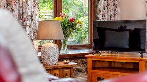 Un salon avec un téléviseur posé sur un meuble en bois, une lampe, un vase de fleurs et une fenêtre avec des rideaux.