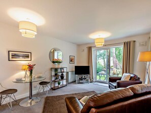 A modern living room with a glass dining table, a leather sofa, a flat-screen TV, and a large window offering a view of greenery. - 1 Bed in Ambleside (oc-94656) (Grasmere)