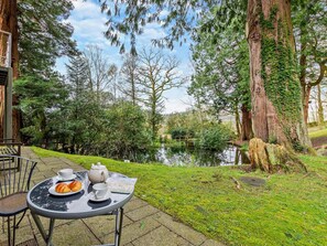 An outdoor seating area with a table set for tea and pastries, surrounded by trees and a body of water. - 1 Bed in Ambleside (oc-94656) (Grasmere)