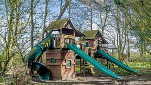 Ein hölzernes Spielhaus mit zwei Rutschen in einem bewaldeten Gebiet.