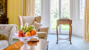 Ein Zimmer mit einer Couch, einem Holztisch mit Früchten, einem Schachbrett und Blick durch große Fenster.