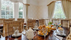 Ein Esszimmer mit Korbstühlen, einem Holztisch, der für das Frühstück gedeckt ist, und großen Fenstern mit Vorhängen.