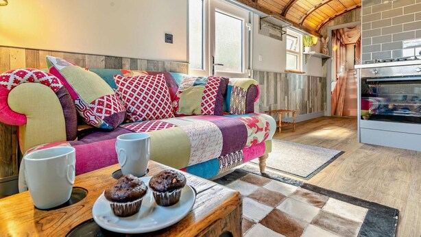 Una zona d'estar acollidora amb sostre de fusta, un sofà amb coixins de colors, una taula de cafè amb muffins i una cuina amb pared de maó.