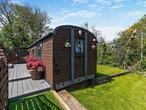 A wooden cabin with a deck, surrounded by a grassy yard and trees. - 1 Bed in Redruth (oc-93858) (Redruth)