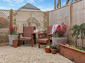 Un área de estar al aire libre con muebles de madera, sillas y una mesa, rodeada de plantas en macetas y una valla de madera.