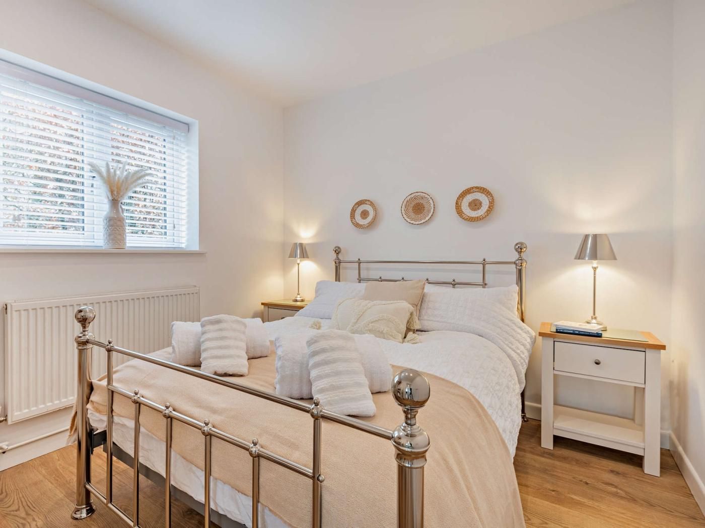 A bedroom with a metal-framed bed, bedside tables with lamps, a radiator, and a window with blinds.