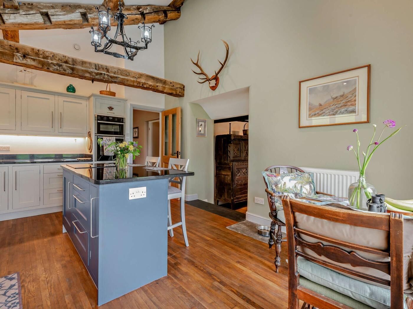 A kitchen with wooden beams, a chandelier, a deer head decoration, and a dining area with chairs and a table.