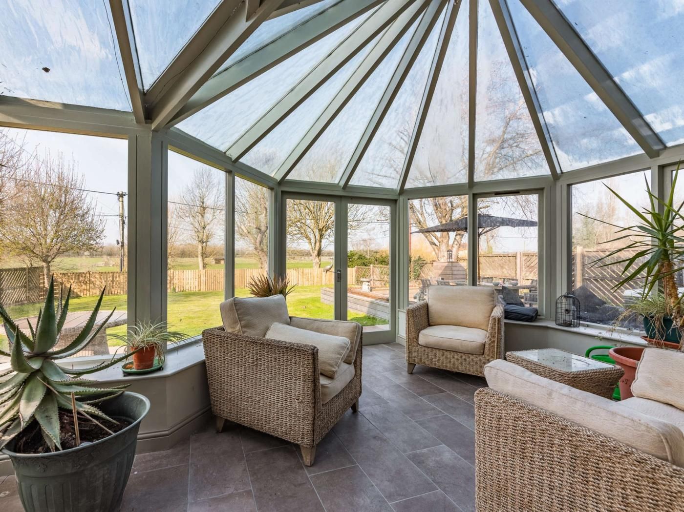 A conservatory with wicker furniture, a glass ceiling, and a view of the garden.