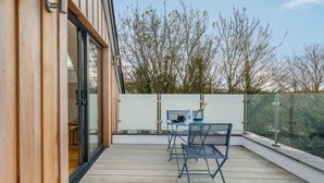 Eine Holzterrasse mit einem Glastisch und Stühlen, einer Champagnerflasche und Blick auf Bäume.